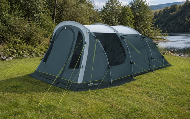 Grijsgroene tunneltent met scheerlijnen op een grasveld aan de oever van een meer; bomen en beboste heuvels op de achtergrond, lucht erboven