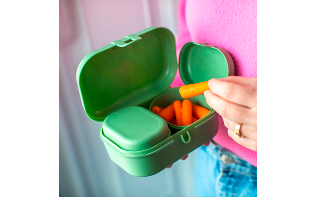 Boîte-repas verte ouverte contenant des mini-carottes ; une main tient une carotte. Personne portant un haut rose et un jean bleu ; arrière-plan flou