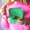 Personne en pull rose tient une boîte plastique verte ouverte contenant des bouchons d’oreille en mousse orange; une main tient un bouchon près de la bouche; seul le bas du visage est visible
