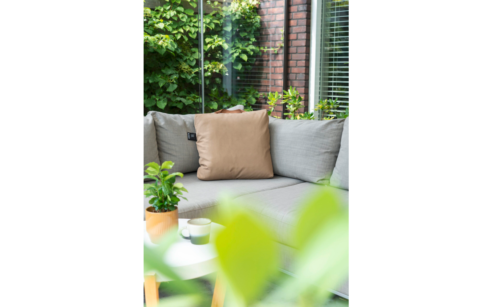 Grijze bank met beige kussen, kleine ronde tafel met plantenpot en kopje, glazen wand, bakstenen muur en groene planten, onscherpe bladeren op de voorgrond