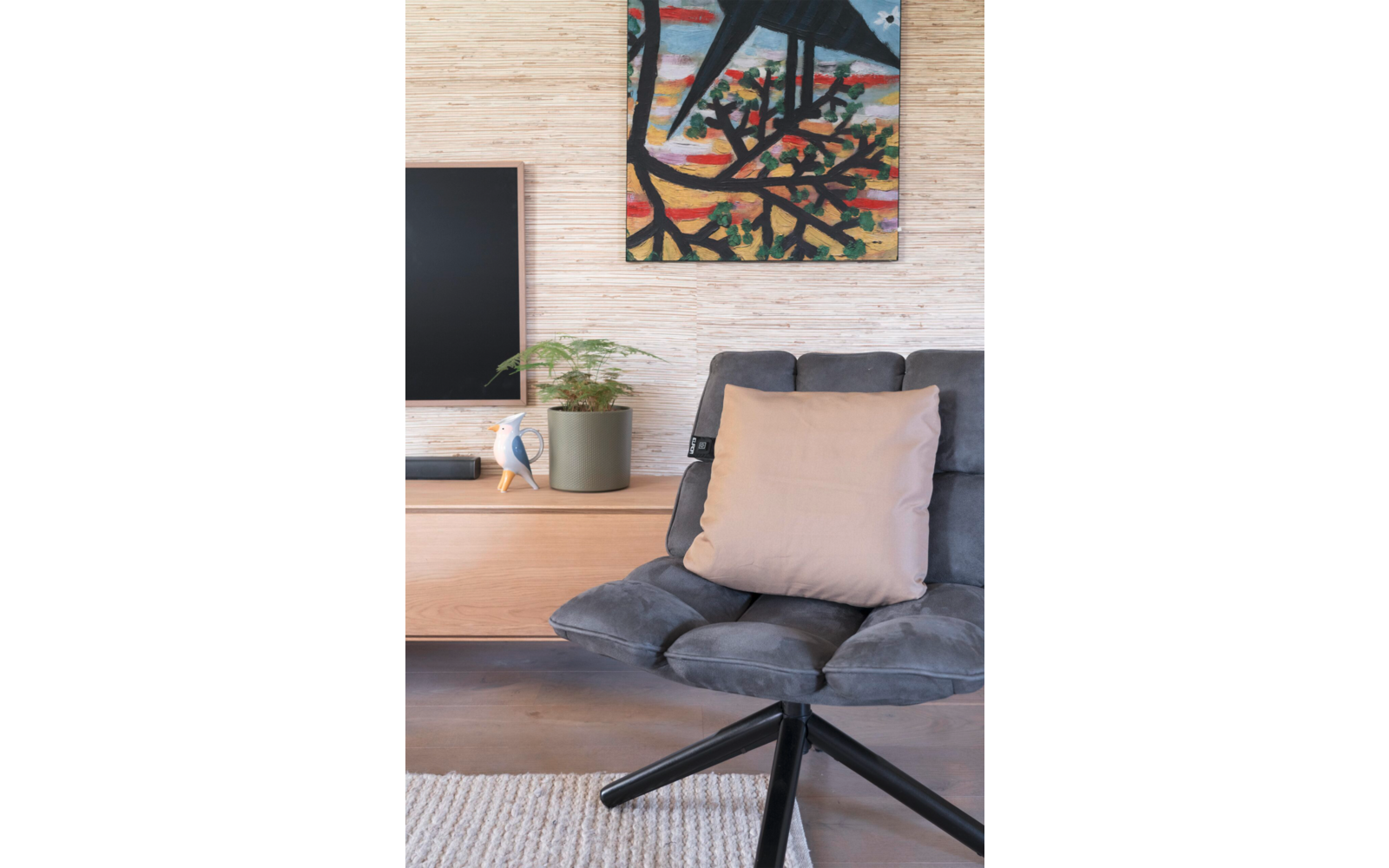 Grijze draaistoel met beige kussen voor een houten dressoir; op het dressoir een kamerplant, een wit decoratief object en een zwarte televisie; lichte muur met een kleurrijk schilderij erboven