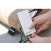 Handen houden een witte powerbank met het 'VARTA'-logo en steken een USB-kabel in; beige tas op de achtergrond