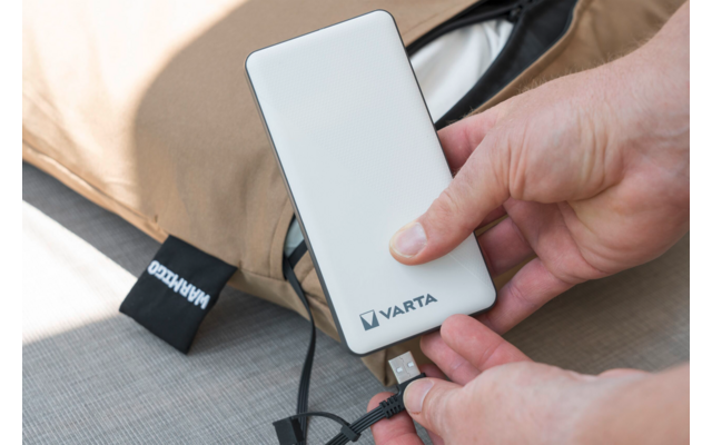 Handen houden een witte powerbank met het 'VARTA'-logo en steken een USB-kabel in; beige tas op de achtergrond