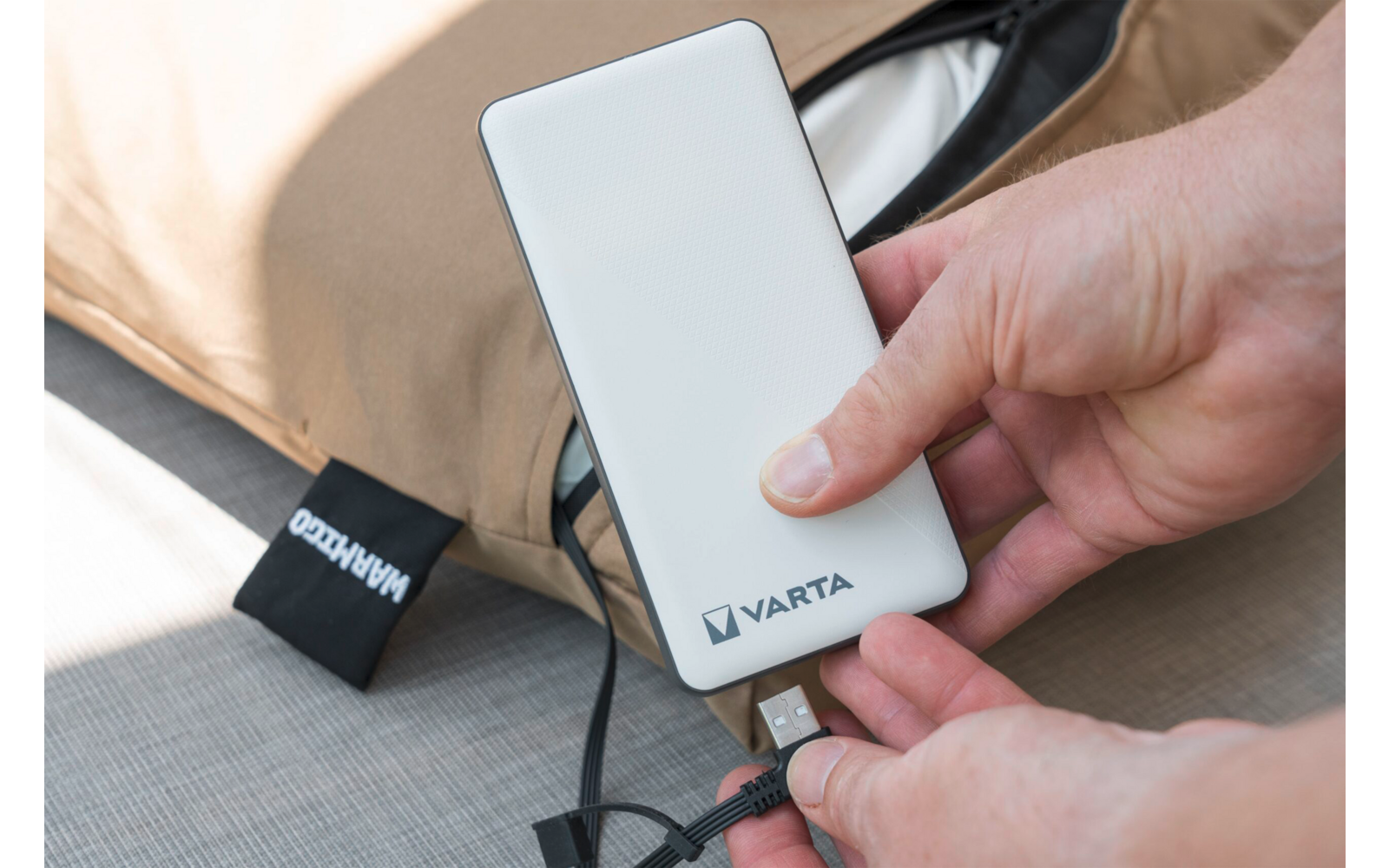 Handen houden een witte powerbank met het 'VARTA'-logo en steken een USB-kabel in; beige tas op de achtergrond