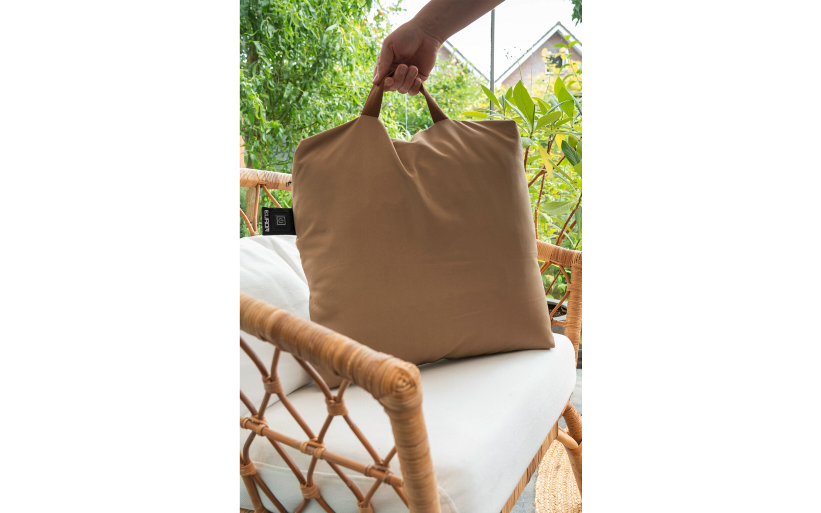 Hand houdt een beige kussen met handgreep vast op een rotanstoel met wit zitkussen, planten en een huisdak op de achtergrond