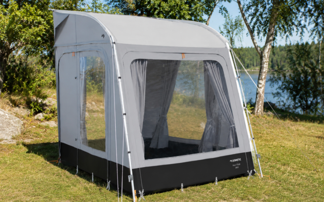 Wit-grijze tent/voortent met grote transparante ramen, zwarte onderrand, scheerlijnen en metalen stangen, op gras bij een meer met bomen op de achtergrond
