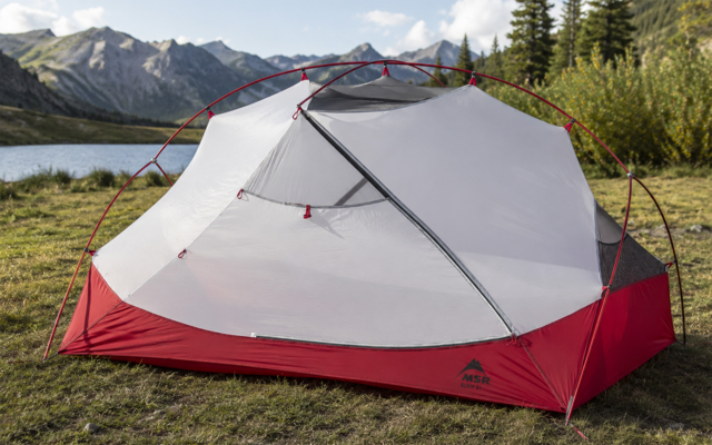 Tente rouge et blanche avec habitacle en moustiquaire et arceaux sur herbe, lac, montagnes, arbres et ciel partiellement nuageux en arrière-plan