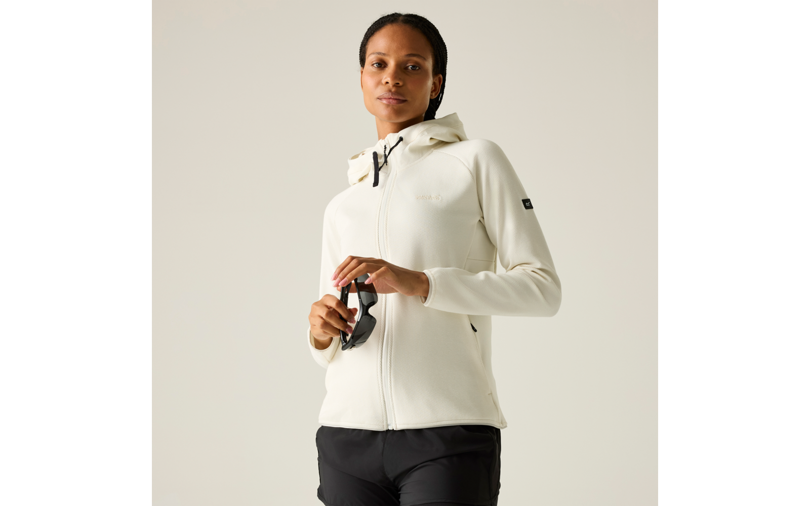 Person in heller Kapuzenjacke und schwarzer Hose, hält schwarze Sonnenbrille vor neutralem hellem Hintergrund