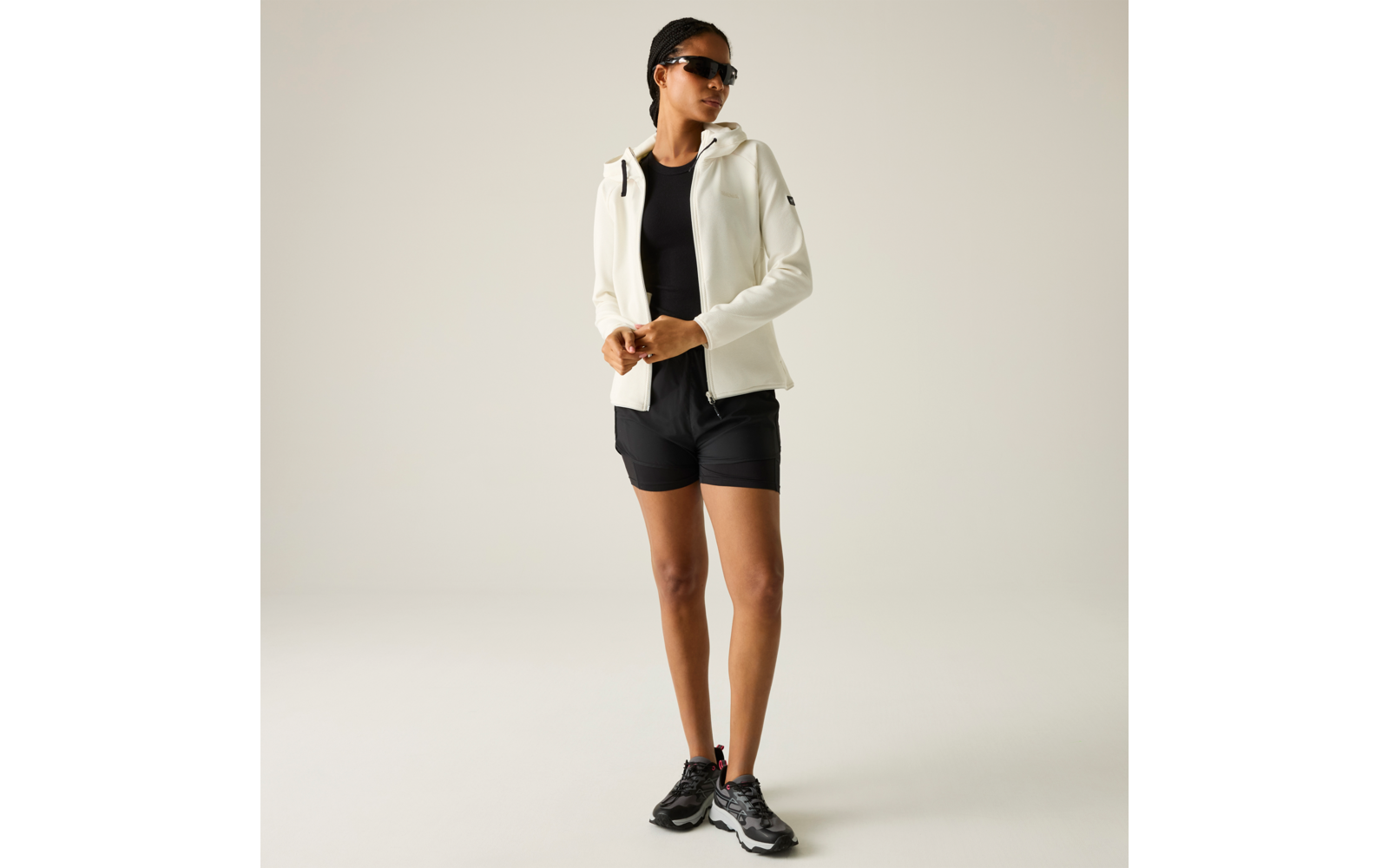 Person mit Sonnenbrille, weißer Jacke, schwarzem Top, schwarzen Shorts und schwarzen Sportschuhen steht vor hellem Hintergrund, die Hände vor dem Bauch gefaltet