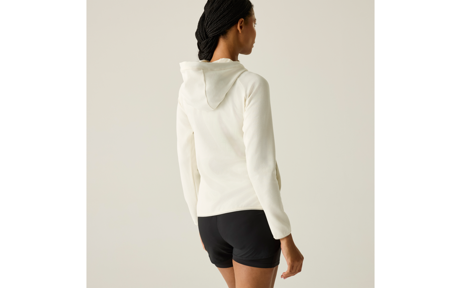 Rückansicht einer stehenden Person mit dunklen, geflochtenen Haaren, weißem Kapuzenpullover und schwarzen Shorts vor hellem Hintergrund