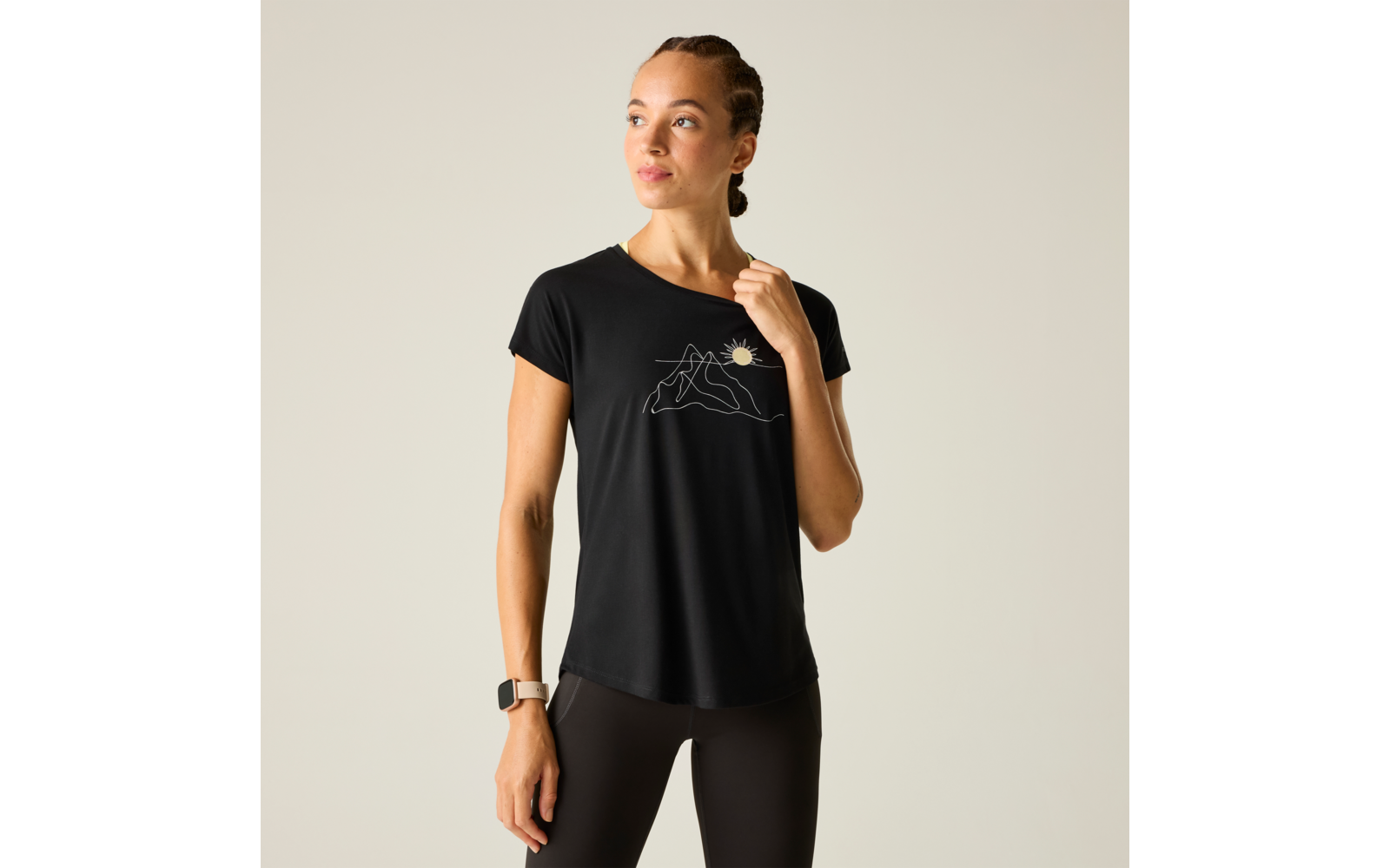 Frau in schwarzem T-Shirt und Leggings vor hellem Hintergrund, Haare geflochten, Armbanduhr am Handgelenk, rechte Hand am Kragen, weiße Linienzeichnung von Bergen und Sonne auf dem T-Shirt