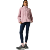 Persoon staand voor een lichte achtergrond, draagt een roze fleecejas met ritssluiting en borstzak, donkerblauwe legging, roze sokken en donkere sportschoenen, haar naar achteren gebonden, één hand in de jaszak