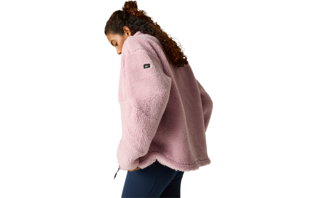 Zijaanzicht van een staande persoon met krullend haar die een roze fleecejas en donkere legging draagt voor een lichte egale achtergrond