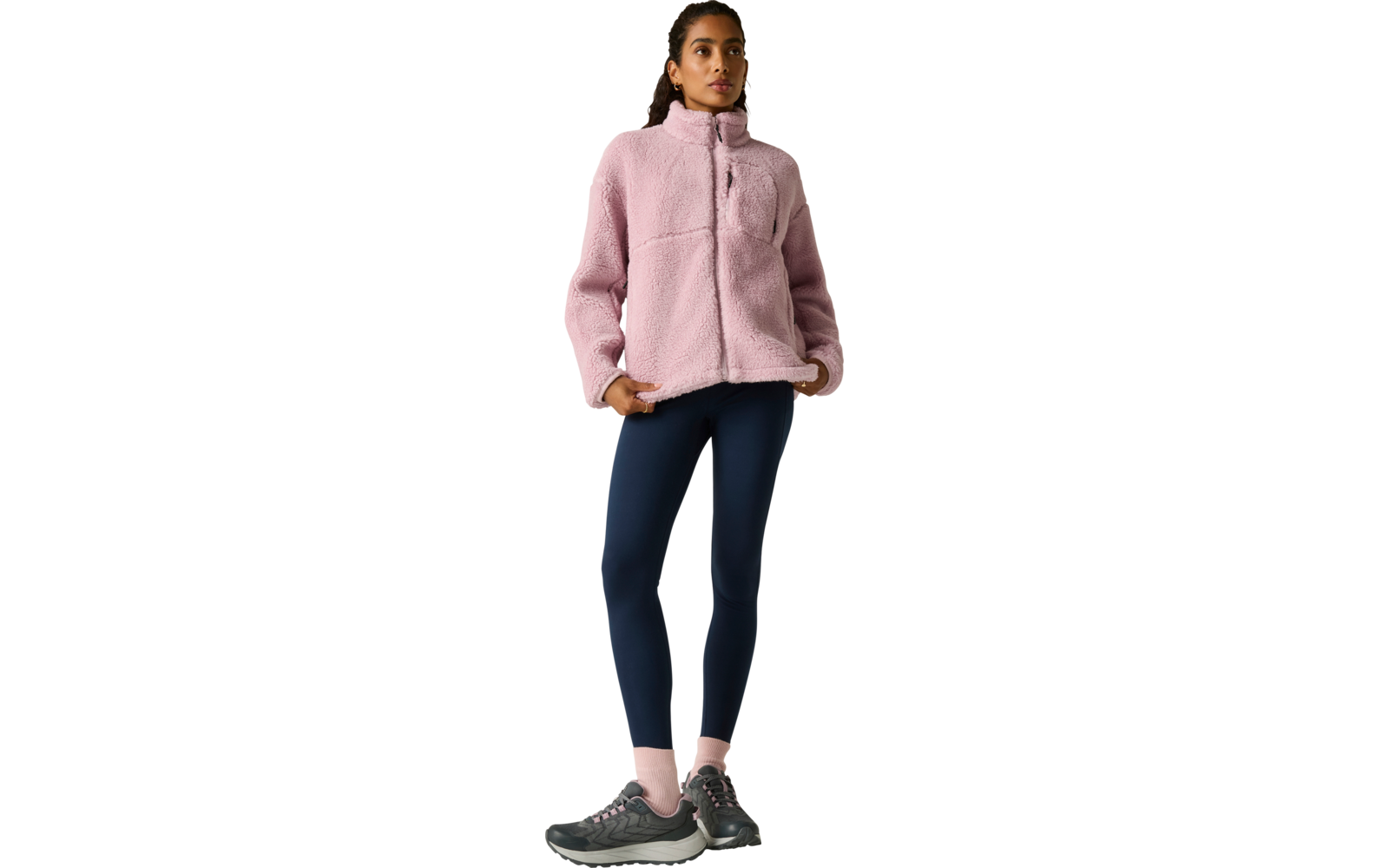 Persoon staand voor een lichte achtergrond, draagt een roze fleecejas met ritssluiting en borstzak, donkerblauwe legging, roze sokken en donkere sportschoenen, haar naar achteren gebonden, één hand in de jaszak
