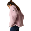 Zijaanzicht van een staande persoon met krullend haar die een roze fleecejas en donkere legging draagt voor een lichte egale achtergrond
