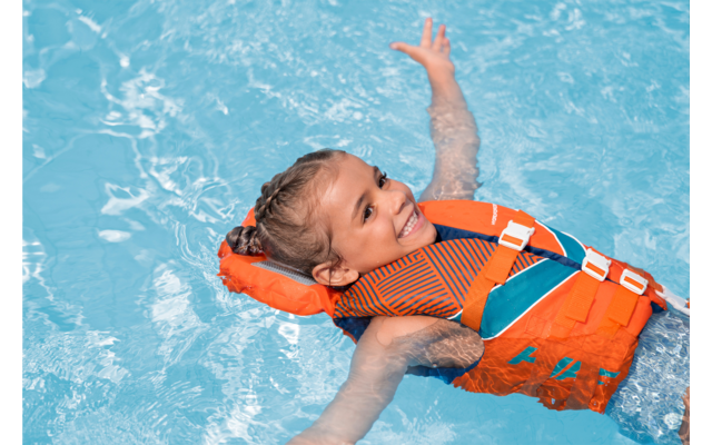 Kind mit geflochtenen Haaren treibt auf dem Rücken in einem blauen Schwimmbecken, trägt eine orangefarbene Schwimmweste, lächelnd, Arme ausgestreckt