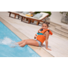 Kind mit orangefarbener Schwimmweste sitzt am Poolrand, Füße im Wasser, zwei Liegestühle im Hintergrund