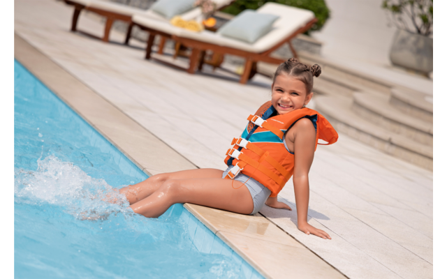Kind mit orangefarbener Schwimmweste sitzt am Poolrand, Füße im Wasser, zwei Liegestühle im Hintergrund