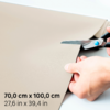 Deux mains coupent un matériau beige avec des ciseaux sur fond blanc ; en coin, dimensions noires 70,0 cm x 100,0 cm et 27,6 in x 39,4 in visibles