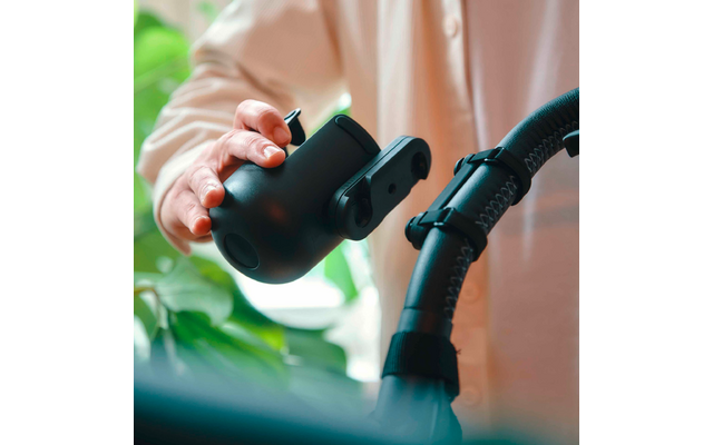 Hand hält ein schwarzes Kunststoffteil neben einem schwarzen Schlauch mit Klemmverschluss; unscharfer Hintergrund mit grünen Pflanzen und hellem Hemd