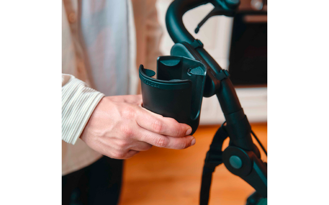 Hand mit hell gestreiftem Ärmel hält einen schwarzen Kunststoff-Becherhalter an einem schwarzen Griff; Holzboden im Hintergrund