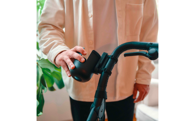 Person von der Brust abwärts in hellbeiger Bluse berührt mit einer Hand einen schwarzen runden Aufsatz an einem schwarzen Rollatorgriff; Pflanzenblatt und Sofa im Hintergrund