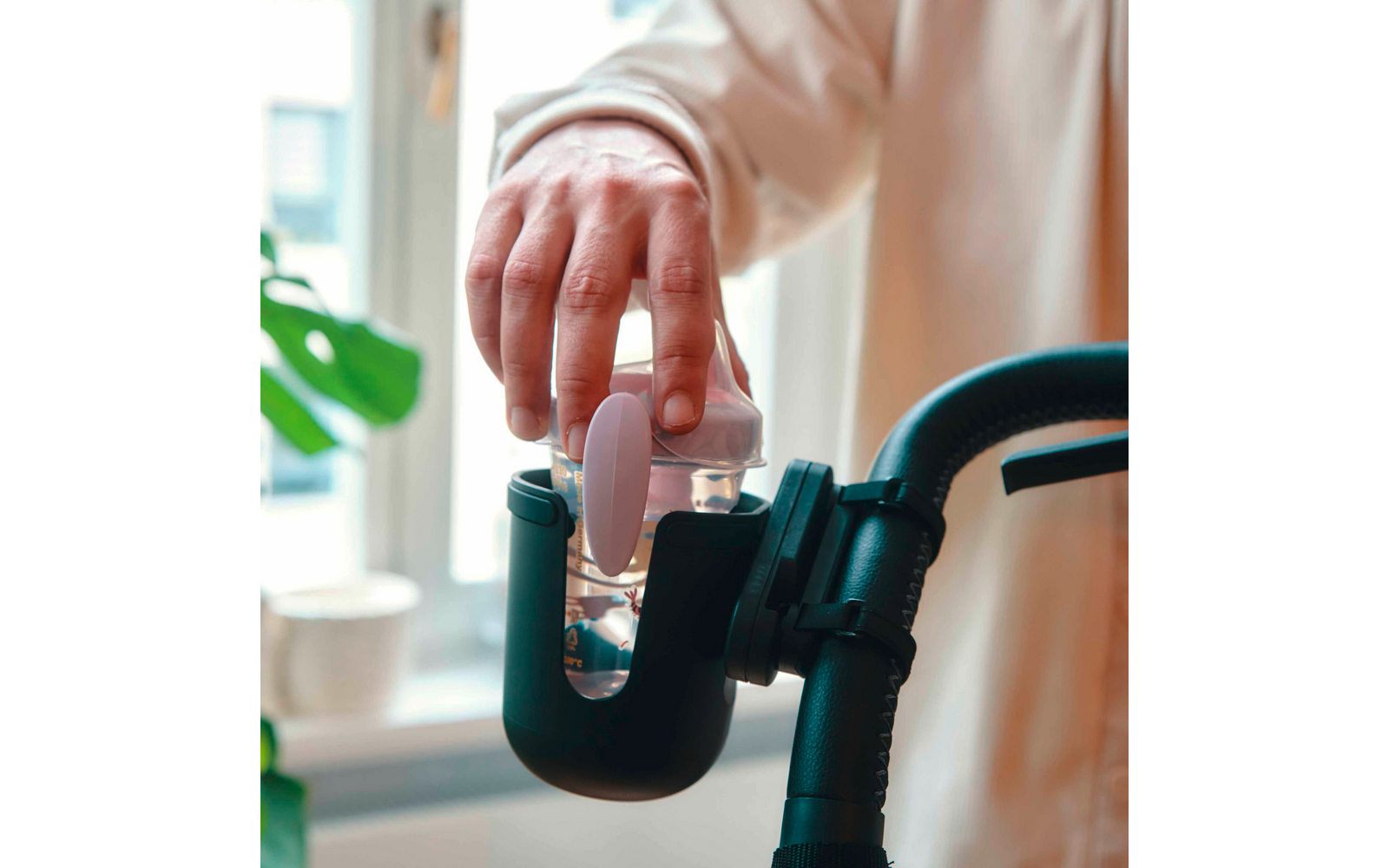 Hand in hellbeigem Ärmel stellt eine durchsichtige Flasche mit Messskala und rosa Sauger in einen schwarzen Halter an einem gebogenen Griff; Fenster und grüne Zimmerpflanze im Hintergrund