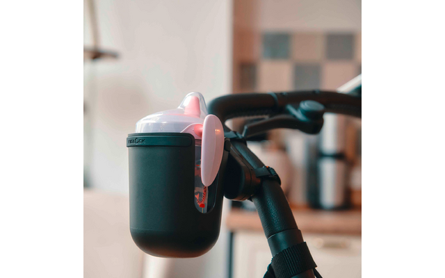 Schwarzer Trinkbecher mit rosa-weißem Deckel, an einem schwarzen Griff befestigt; unscharfer Hintergrund mit Küchenfliesen und Geräten