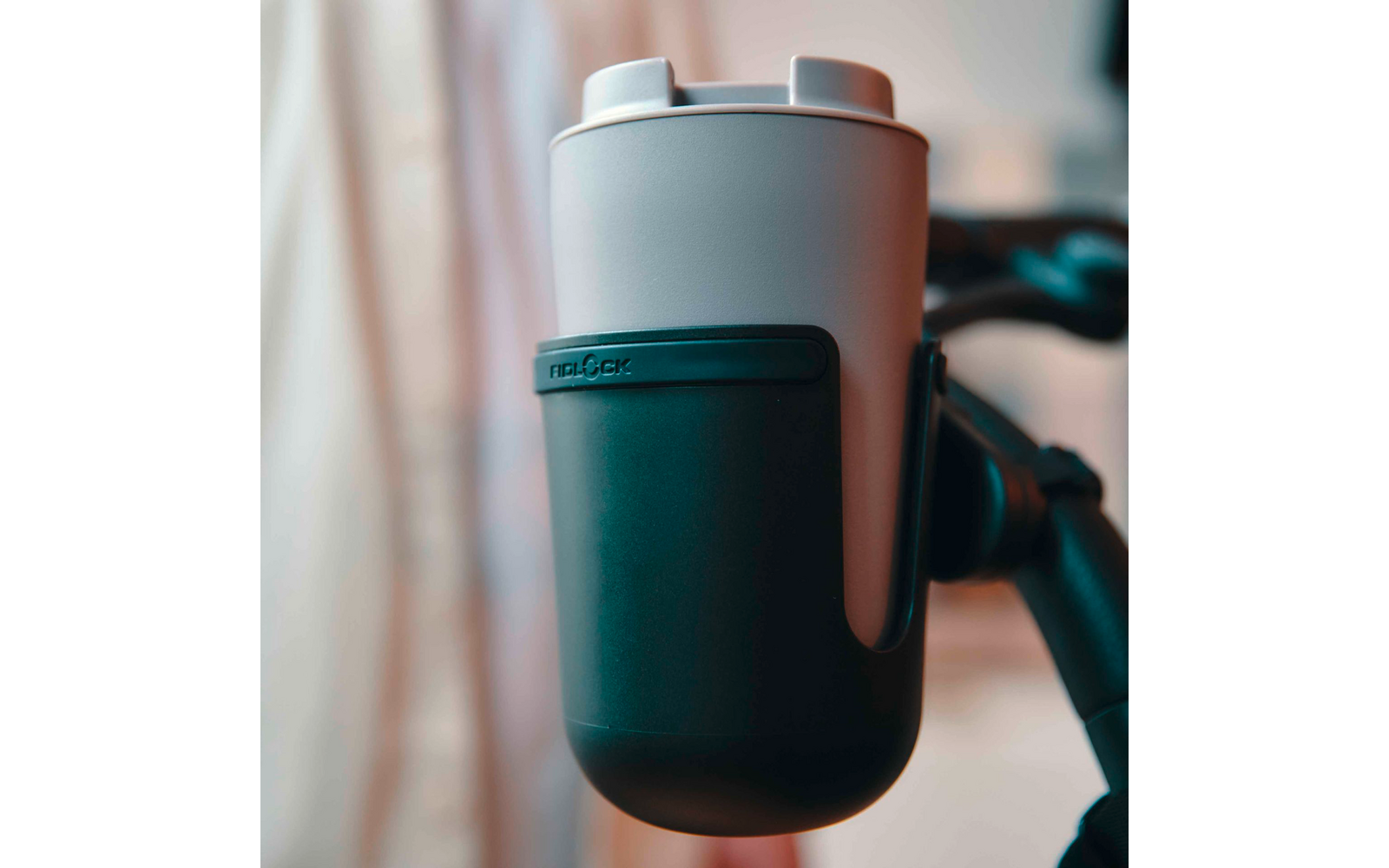 Hellgrauer zylindrischer Becher mit hellem Deckel und schwarzer Silikonhülle in schwarzem Halter; unscharfer heller Vorhang im Hintergrund