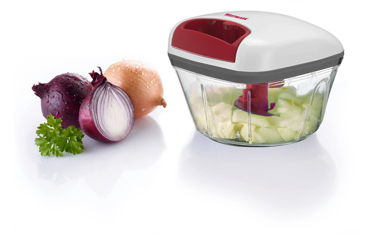 Petit hachoir de cuisine avec couvercle blanc et rouge et récipient transparent contenant des morceaux verts hachés ; à côté, un oignon rouge coupé en deux, un oignon brun entier et un brin de persil