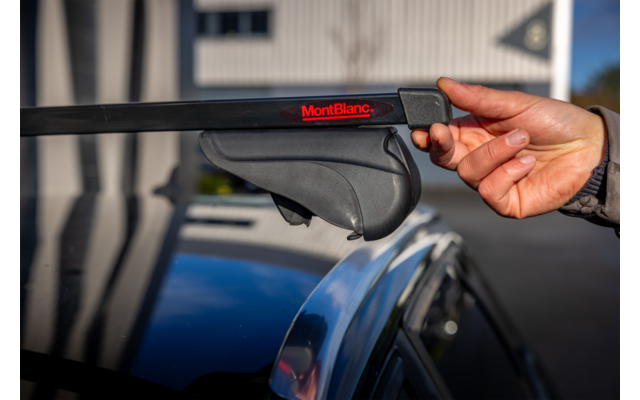 Hand hält schwarzen Dachträger-Querträger mit rotem 'MontBlanc'-Logo über silberner Dachreling eines Autos; unscharfes Gebäude im Hintergrund