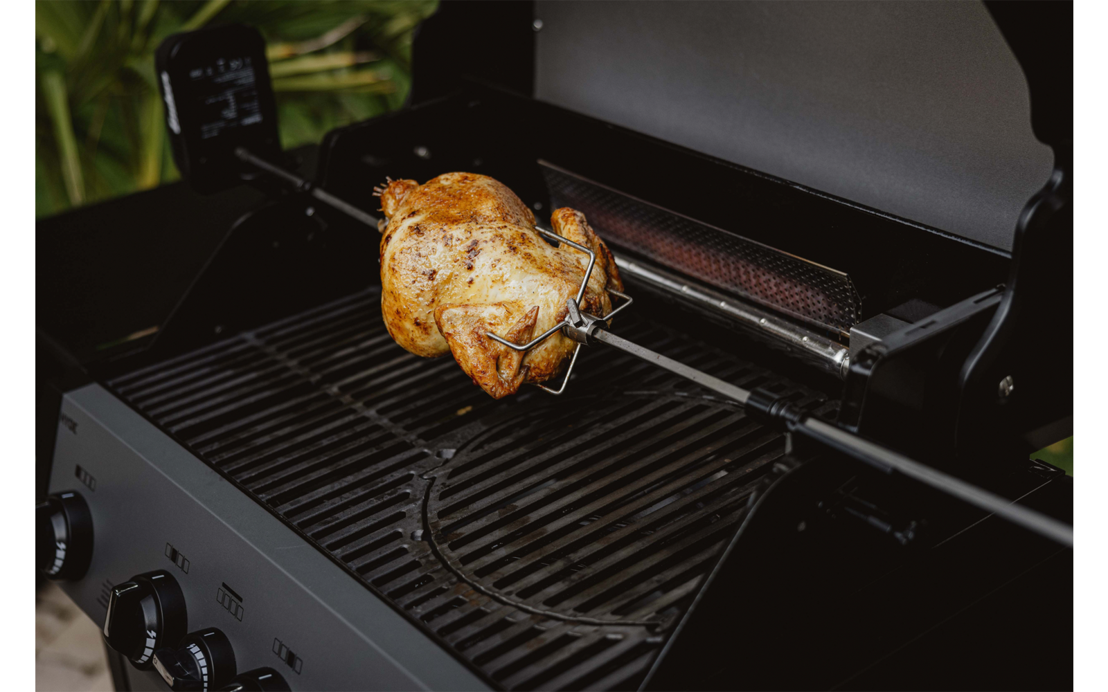 Ganzes Hähnchen auf Drehspieß über einem Gasgrill, Grillrost und Bedienelemente sichtbar