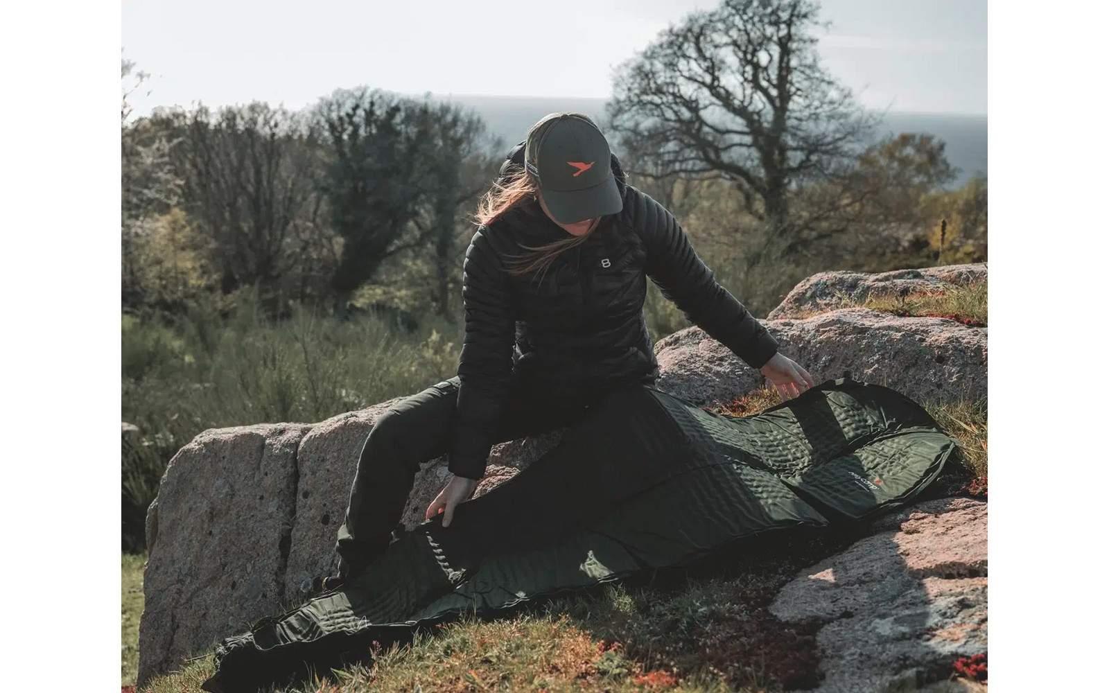 Person mit schwarzer Kleidung und Mütze sitzt auf einem großen Stein in einer natürlichen Umgebung mit Bäumen und Sträuchern