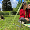 Zwei Personen sitzen auf einer Decke im Gras; eine trägt einen roten Pullover und blickt nach rechts, die andere ist teilweise sichtbar. Links liegt ein Hund auf dem Rasen. Im Hintergrund Hecke, hohe Bäume und teils wolkiger blauer Himmel. Grashalme 