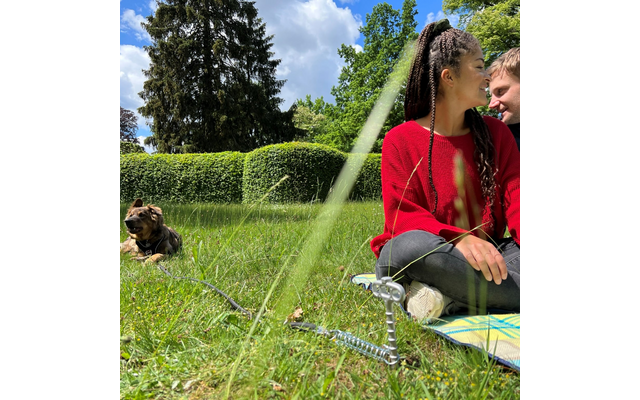 Zwei Personen sitzen auf einer Decke im Gras; eine trägt einen roten Pullover und blickt nach rechts, die andere ist teilweise sichtbar. Links liegt ein Hund auf dem Rasen. Im Hintergrund Hecke, hohe Bäume und teils wolkiger blauer Himmel. Grashalme 