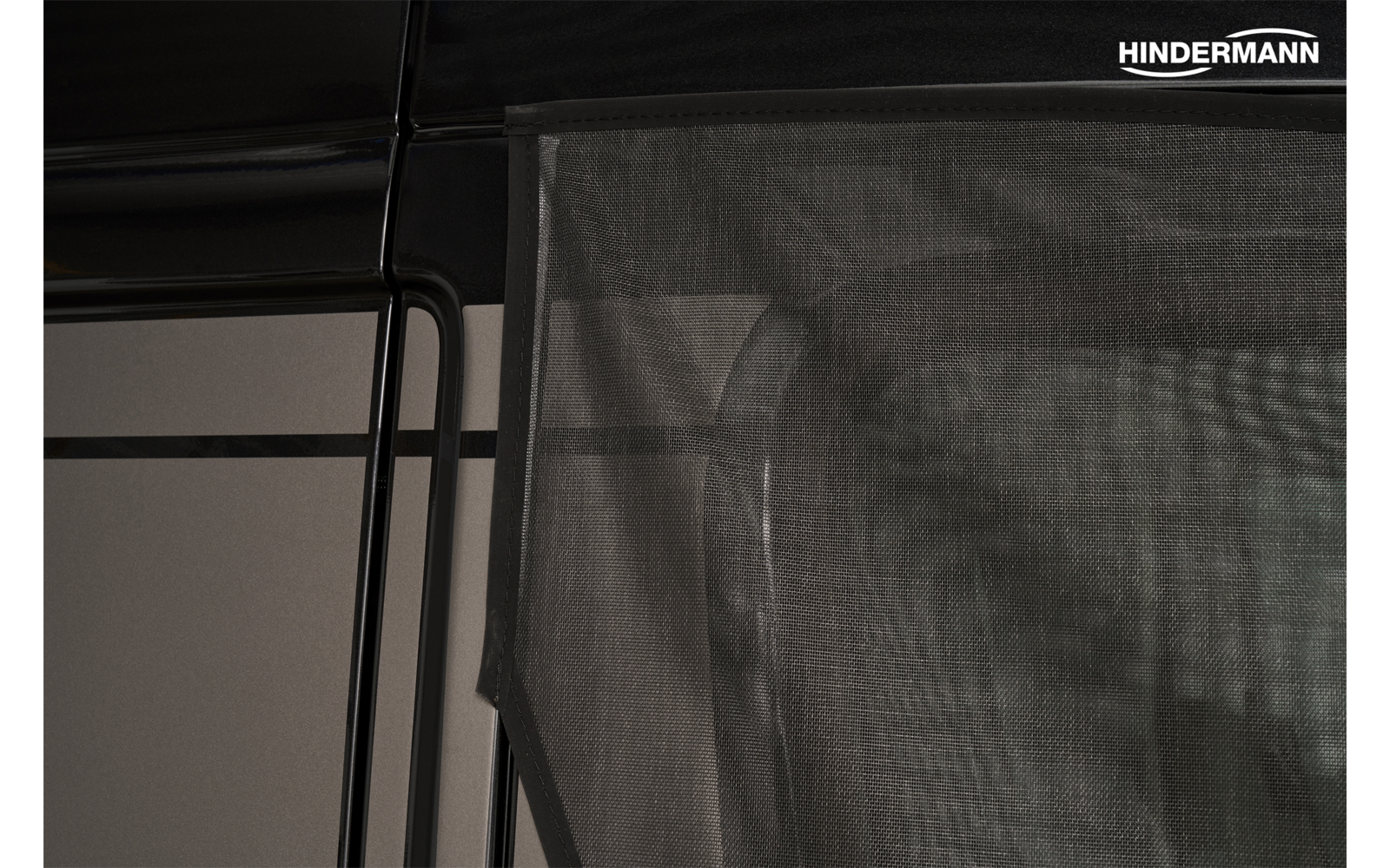 Tenda a rete scura semitrasparente davanti a un'apertura rettangolare con telaio metallico nero; scritta bianca 'HINDERMANN' in alto a destra
