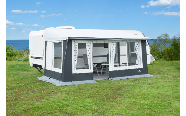 Weißer Wohnwagen mit grauem Vorzelt auf einer grünen Wiese, Meer im Hintergrund und blauer Himmel mit wenigen Wolken; keine Personen sichtbar