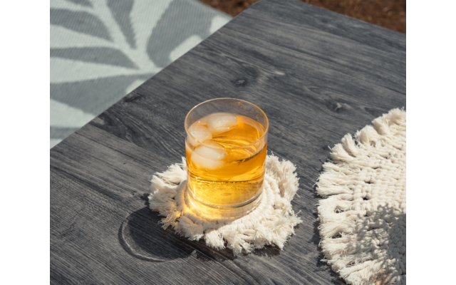 Verre transparent contenant un liquide ambré et des glaçons sur une table en bois foncé, posé sur un dessous de verre blanc à franges ; un autre dessous de verre à droite et un tapis à motifs à gauche