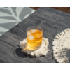 Verre transparent contenant un liquide ambré et des glaçons sur une table en bois foncé, posé sur un dessous de verre blanc à franges ; un autre dessous de verre à droite et un tapis à motifs à gauche