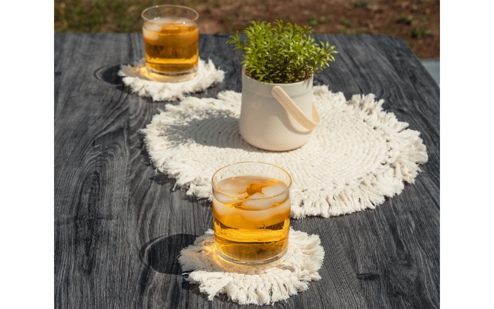 Deux verres avec une boisson dorée et des glaçons, un petit pot avec une plante verte et des sets blancs en crochet sur une table en bois foncé