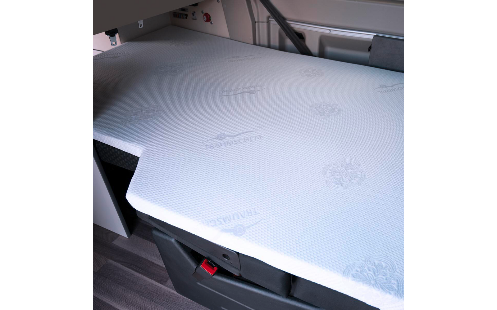 Weißes, strukturiertes Matratzenpolster mit eingeprägten Logos auf einer zugeschnittenen Liegefläche; darunter dunkles Staufach mit rotem Verschluss, Laminatboden und seitliche Innenverkleidung