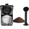 Aufgeklappte schwarze Kaffeemaschine mit sichtbarer silberner Kapselkammer; daneben ein Haufen Kaffeepulver und ein schwarzer Kunststofflöffel mit langem Stiel
