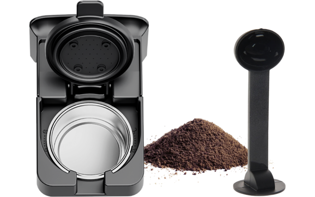 Aufgeklappte schwarze Kaffeemaschine mit sichtbarer silberner Kapselkammer; daneben ein Haufen Kaffeepulver und ein schwarzer Kunststofflöffel mit langem Stiel