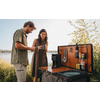 Ein Mann und eine Frau stehen an einem mobilen Camping-Küchenmodul am Ufer und halten Tassen; auf der Arbeitsfläche stehen eine Kaffeemaschine, Küchenutensilien und Vorratsdosen