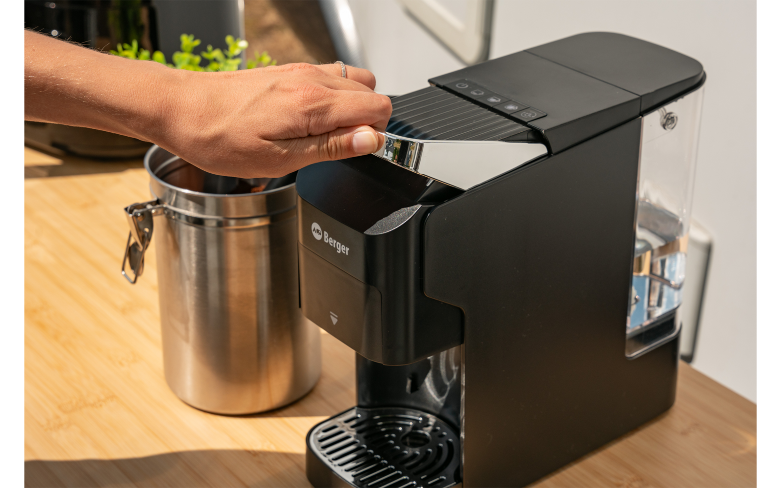 Zwart koffieapparaat op houten aanrecht, transparant waterreservoir aan de zijkant, hand tilt een zilveren hendel op, roestvrijstalen melkkan naast het apparaat, kleine groene plant op de achtergrond