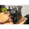 Hand trekt de voorste inzet van een zwarte koffiemachine, apparaat staat op een licht houten aanrecht; links een potplant en een metalen bak