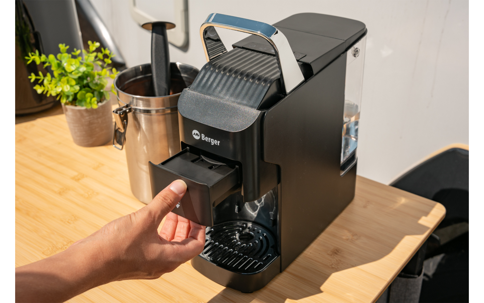 Hand trekt de voorste inzet van een zwarte koffiemachine, apparaat staat op een licht houten aanrecht; links een potplant en een metalen bak
