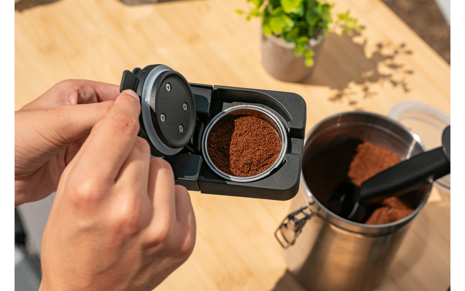 Twee handen houden een zwarte koffiepadhouder met open deksel; gemalen koffie ligt erin. Rechts een metalen bus met gemalen koffie en een zwarte schep. Licht houten tafel en een kleine potplant op de achtergrond