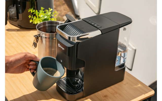 Compact zwart koffiezetapparaat op houten aanrecht, metalen melkkan ernaast, een hand houdt een grijze mok onder de schenktuit