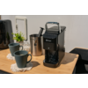 Zwarte capsulekoffiemachine op houten aanrecht, twee donkere mokken op onderzetters, roestvrijstalen bak met capsuleschep, glazen pot met deksel, kleine potplant